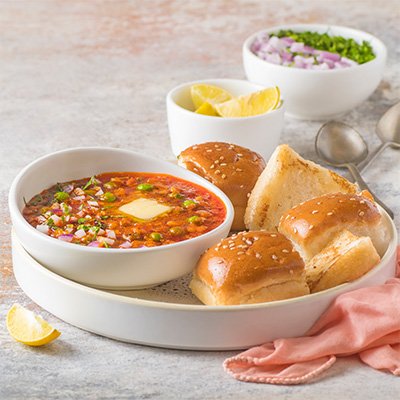 A serving of Pav Bhaji, featuring a rich and spicy vegetable mash served with buttered and toasted pav (bread rolls), garnished with chopped onions, cilantro, and a wedge of lime.