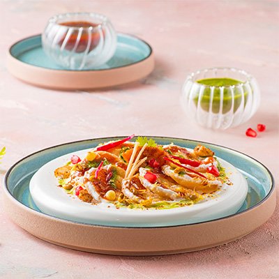 A vibrant plate of Papdi Chaat, showcasing crispy papdis topped with spiced potatoes, chickpeas, creamy yogurt, tangy tamarind chutney, and garnished with fresh cilantro and sev.