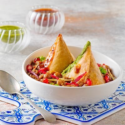 A colorful plate of Samosa Chaat, featuring crispy samosas topped with tangy yogurt, spicy chutneys, chopped onions, and fresh cilantro, garnished with sev for added crunch.